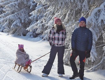 Junge Familie stehen in einem winterlichen Wald, Tochter wird auf Schlitten gezogen