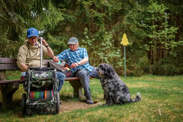Zwei Männer mit Hund machen eine Pause vom DiscGolf und ruhen sich auf einer Bank aus
