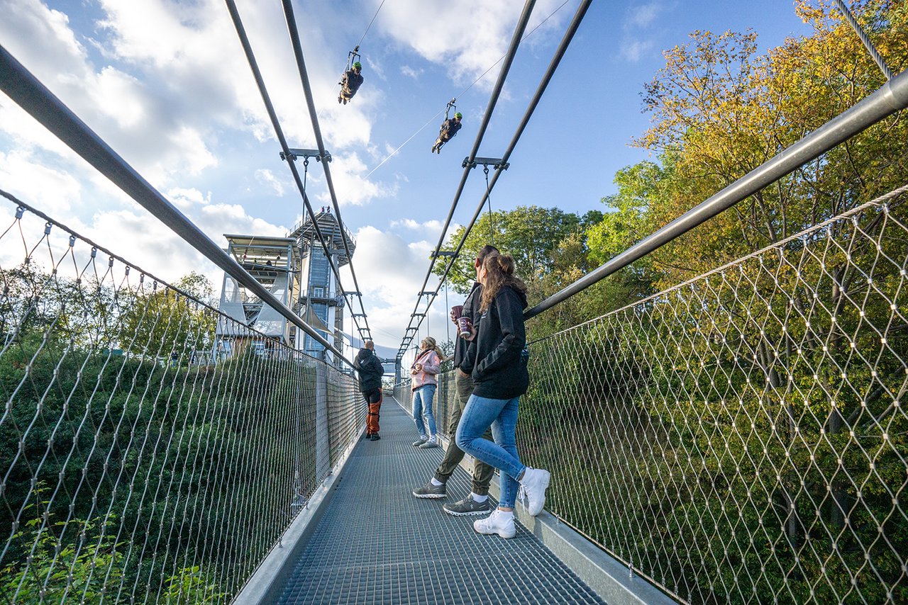 Personen stehen auf Hängebrücke und schauen auf vorbeirasende Personen an Zipline 