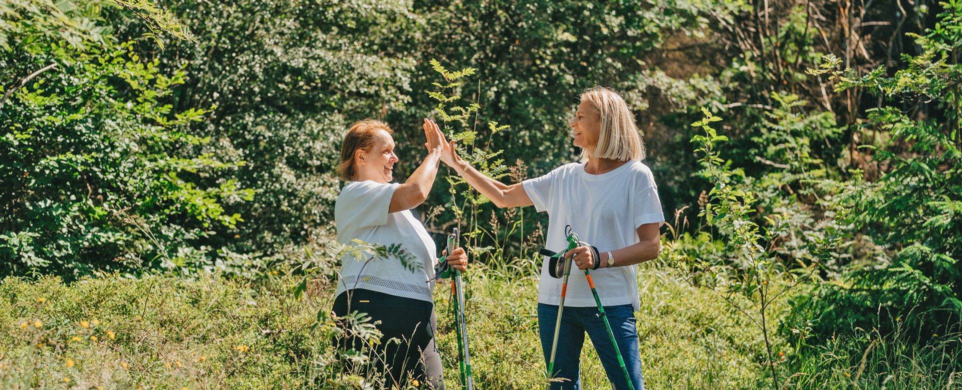 Zwei Frauen mit Nordic-Walking Stöcken stehen im Wald in geben sich High-Five