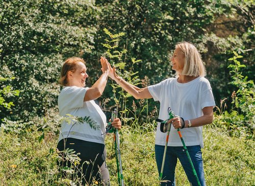 Zwei Frauen mit Nordic-Walking Stöcken stehen im Wald in geben sich High-Five