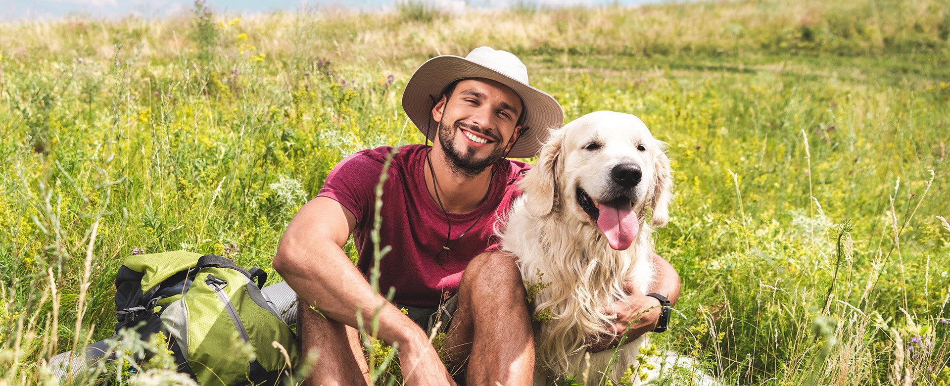 Mann mit Hund sitzen gemütlich auf einer hohen Wiese und lächeln in die Kamera