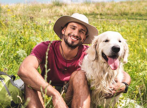 Mann mit Hund sitzen gemütlich auf einer hohen Wiese und lächeln in die Kamera