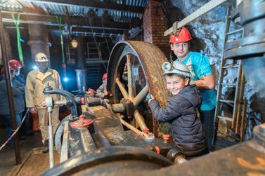 Eine Familie steht an einem alten Förderrad im Stollen und der Stollenführer erläutert die Funktion