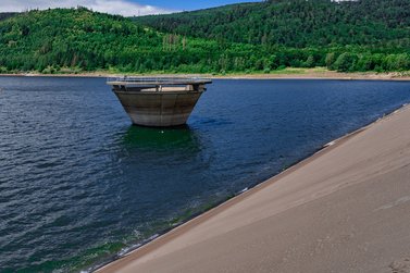 Betonbauwerk ragt aus der Wasseroberfläche einer Talsperre, umgeben von bewaldeten Hängen.