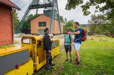 Der Zugführer der Otti-Bahn erklärt zwei Gästen am Ottiliae-Schacht den historischen Bergbau dort 