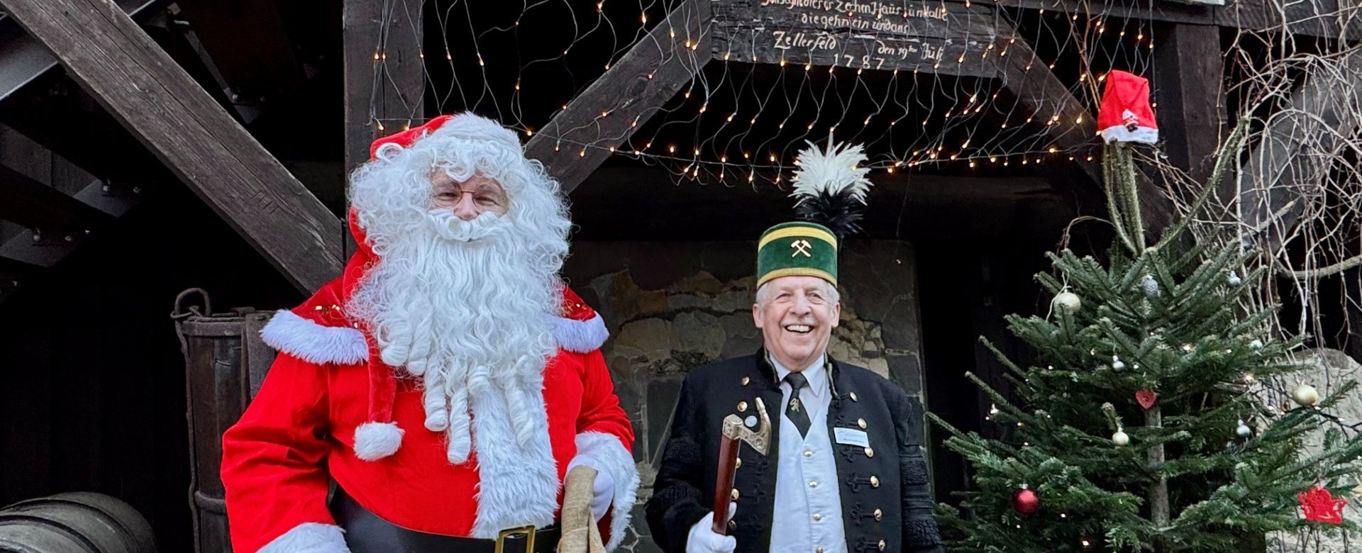 Weihnachtsmann und Bergmann stehen neben Tannenbaum und schauen in Kamera